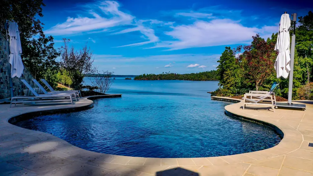Freeform pool with an infinity edge spilling toward a lake under a blue sky, umbrellas and loungers on the deck
