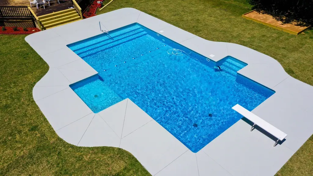 Aerial view of an L-shaped pool with a diving board, white concrete deck and fresh lawn